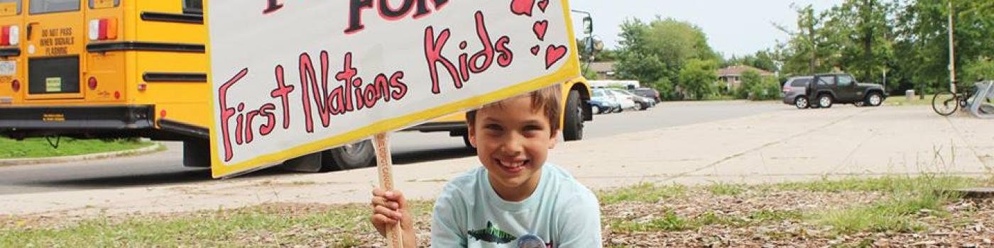 Young boy sitting on the ground holding a sign that reads "Equal Funding for First Nations Kids"